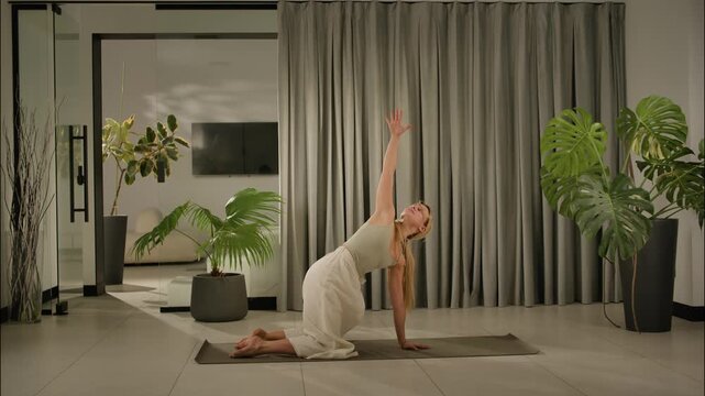 Blonde woman holds kneeling side stretch yoga pose in modern home interior with tropical plants yoga