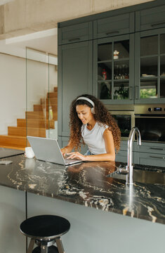 Woman Wearing Headphones Using Laptop In Kitchen