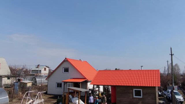 Snow melts beside a metal roofline. Clear skies frame a rustic home. Children play in a yard with friends. Sunlight warms the earth after winter. Family gathering evokes warmth and joy