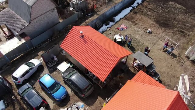 Aerial view of a red-roofed shed surrounded by cars and workers. People move around, possibly repairing or building something. The scene suggests active construction or maintenance work