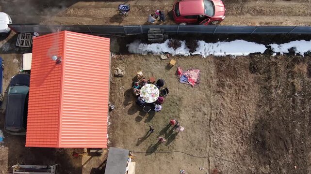 Aerial view of a rural gathering in early spring. Family enjoys a meal outdoors on a round table. Snow remnants linger near the fence. Cars parked nearby, including a blue and a red