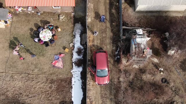 Aerial view of a backyard with a red car and a group of people. Snow melts along a narrow path between two yards. People gather around a table, enjoying a meal