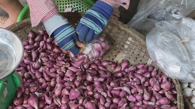 Vendor adding garlic to a bag of shallots on a scale, tying it, and scooping another batch of shallots for weighing at a traditional market