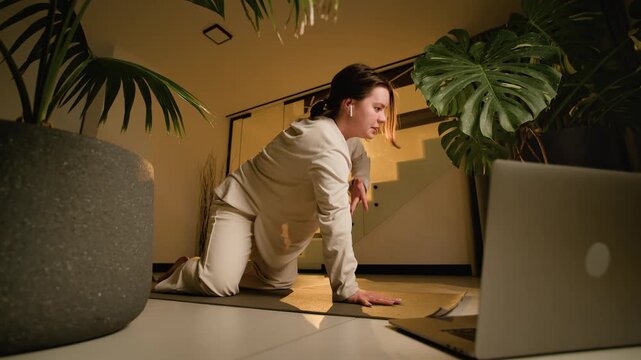 Child pose home yoga stretch with woman resting on forearms beside laptop in warm tropical room pose