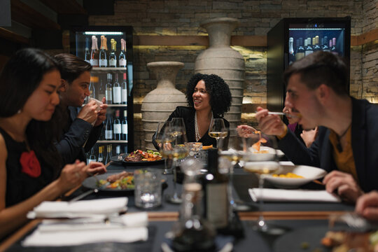 A group of diverse friends enjoys food and drinks while having lively conversations at a restaurant during the evening.