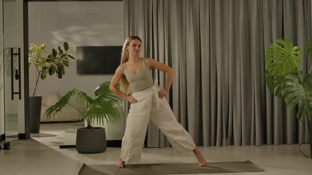 female yoga practitioner performing downward dog in linen set with plants and curtains in side view.