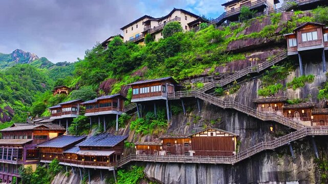 Traditional wooden buildings and stairways cling to a lush rocky cliff, with tiled roofs, dense vegetation, and soft overcast light.