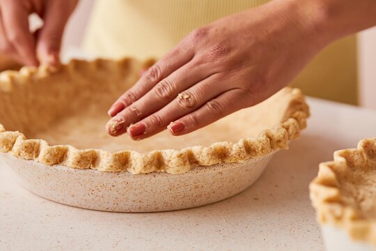 Female hands crimping the edge of a pie crust in a kitchen setting with a second pie crust visible on a countertop, showcasing baking preparation techniques