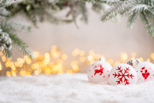 A Christmas background featuring white baubles hand-painted with a red reindeer, snowflake, and candy cane beneath frosted pine on snow, softly lit by warm twinkling lights with copy space.