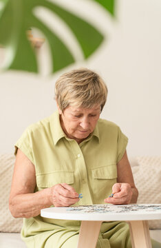 Elderly woman sits on sofa at table, intently working on puzzle, engaging in leisure activities for mental stimulation and brain health. A bright, cozy home interior