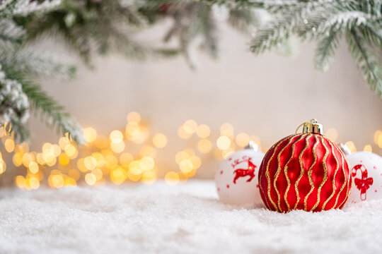 A Christmas background featuring a red plaid snowflake bauble beside white ornaments painted with a reindeer and candy cane beneath frosted pine on snow, softly lit by warm twinkling lights.
