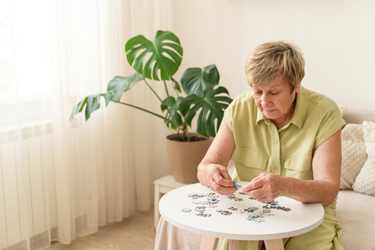 Senior woman actively engages her mind by putting together a puzzle on small table. Slow, mindful living, analog hobby, relaxing activity at home