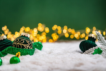 Naklejka premium A Christmas background featuring emerald baubles with gold filigree beneath a frosted pine branch, wrapped in glittering green ribbon on snow, set against deep green with twinkling lights.