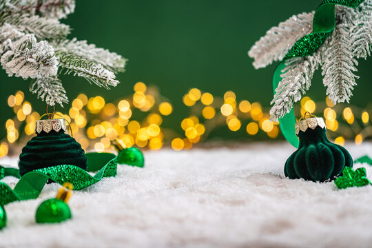 A Christmas background featuring emerald baubles with gold filigree beneath a frosted pine branch, wrapped in glittering green ribbon on snow, set against deep green with twinkling lights.