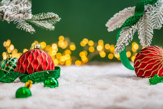 A Christmas background featuring an abundant array of red and green baubles with golden patterns beneath frosted pine branches, wrapped in ribbon on snow, set against deep green with lights.