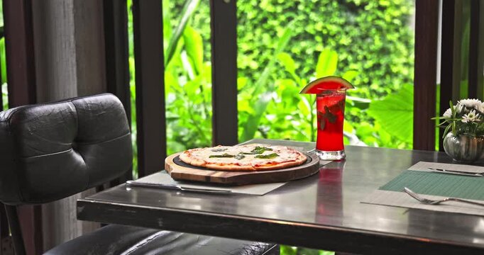 Margherita pizza on wooden board and red watermelon cocktail served on restaurant table. Lush green garden background through window. Summer dining lifestyle, nobody.