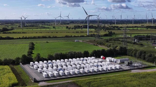 Aerial footage of battery storage facility BESS providing grid stabilization, frequency regulation and power reserves next to wind farm and high-voltage substation.