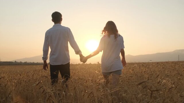 A young couple in love walks hand in hand through a field of wheat or rye toward the sunset, feeling happy and confident in each other. Trust, inner peace, harmony, shared joy.