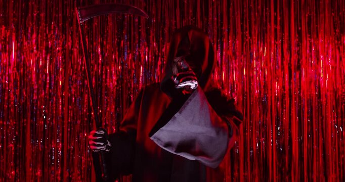 Person in grim reaper death costume saying hush be quiet with fingers on lips with shhh gesture holding scythe and looking at camera on dark red background. Halloween party concept. 4k video.