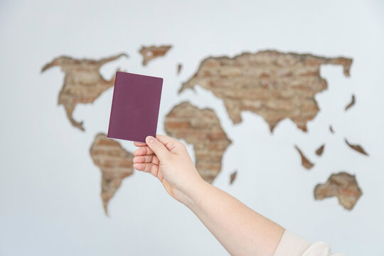 Hand holding a blank burgundy passport cover against a blurred carved world map on a wall