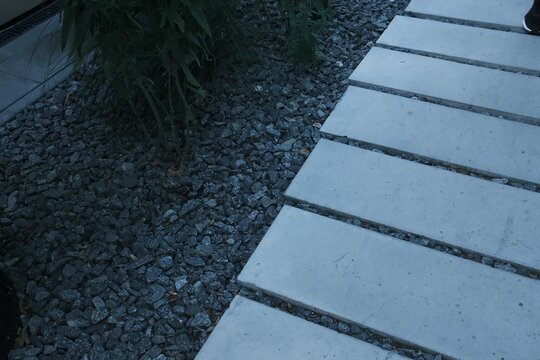 Rectangular light concrete pavers sit next to dark grey decorative gravel in a garden setting during the late evening hours as someone walks along the stone path surrounded by small green plants