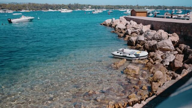 Clear turquoise water ripples along a rocky shoreline in the L&eacute;rins Islands near Cannes, France, with an inflatable dinghy beside a stone jetty and boats anchored offshore.