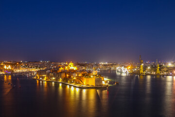 Fototapeta premium Night view of Fort St. Angelo and Birgu town illuminated over the Grand Harbour from the Upper Barrakka Gardens in Valletta, Malta