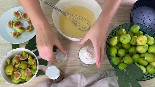 A person handles ingredients for baking, pouring white powder from a glass into a bowl with yellow liquid, surrounded by green figs and sliced figs