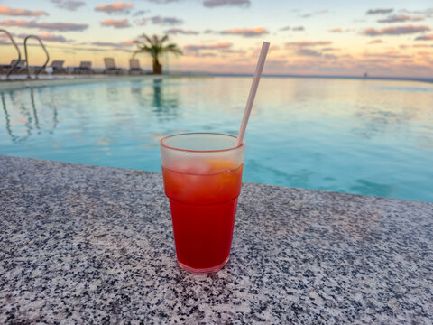Cocktail glass by rooftop pool at sunset, red drink with straw on poolside ledge, tropical vacation relaxation concept for tourism and leisure