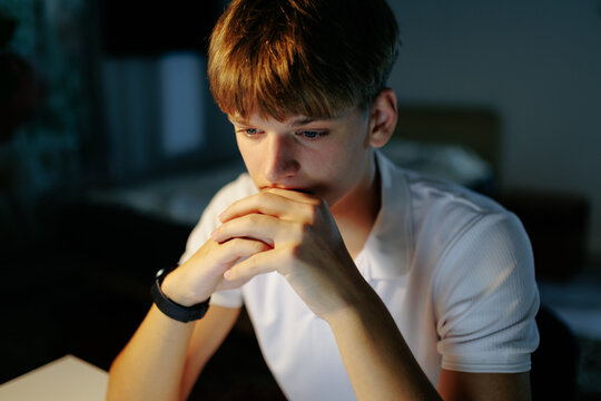 Thoughtful teenage schoolboy looking at a screen doing night homework.