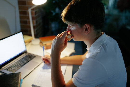 Teenage schoolboy eating a sandwich while writing digital homework at night.