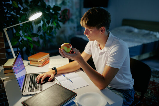 Focused adolescent schoolboy holding an apple doing digital homework at night.