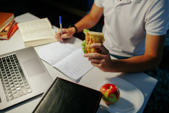 Unrecognizable male student eating food and doing academic homework.
