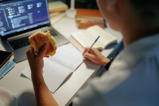 Rear view of a teen schoolboy eating food and writing academic notes.