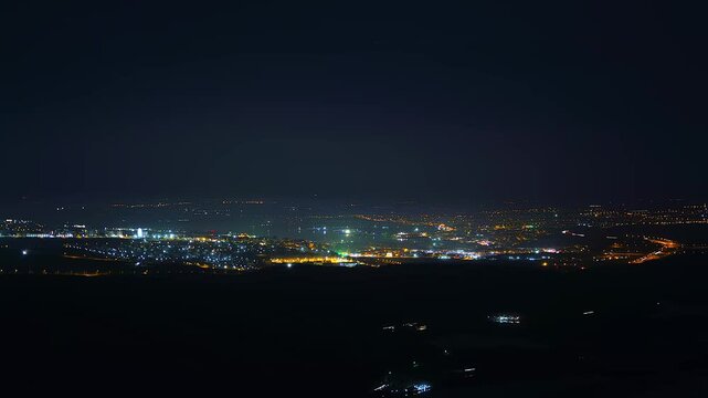Elevated panoramic view of coastal city lights glowing along shoreline at night, warm building windows and streetlights reflecting over dark sea under deep navy sky