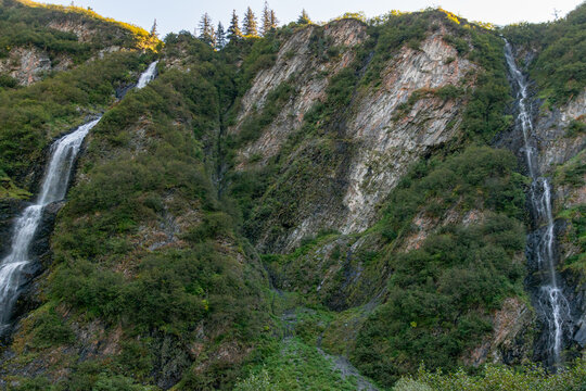 Two waterfalls from mountains on side of winding road in canyon