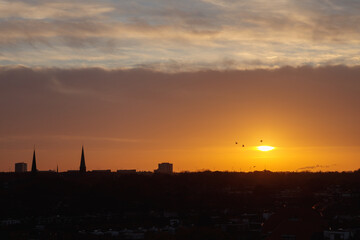 Fiery sunset over silhouetted city skyline with two church spires, tall block, orange and gold sky, three birds in silhouette near sun © Ostrovskajah