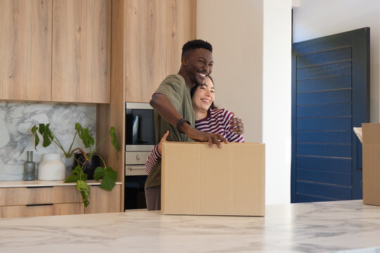 Diverse couple standing behind cardboard moving box on marble kitchen island, embracing and smiling