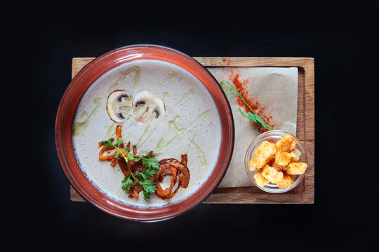 Creamy mushroom soup in ceramic bowl. Garnished with parsley, oil, pepper. Croutons in small glass bowl. Wooden board. Healthy meal. Black background. Top view