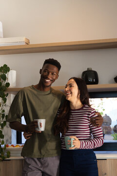 Couple holding ceramic mugs and laughing while leaning on wooden countertop in home kitchen
