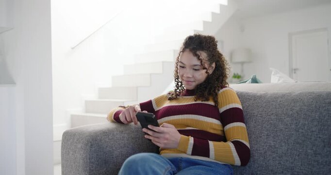 African American female youth sitting on grey sofa, scrolling phone, seeing funny content, smiling
