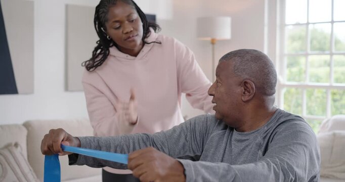African American woman guiding Senior man gripping blue resistance band in living room for strength