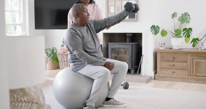 African American aide handing hex dumbbell, guiding senior man lifting on ball at home for strength