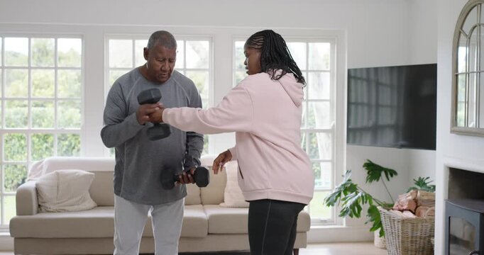 African American daughter in pink hoodie handing hex dumbbells to father, coaching curls at window