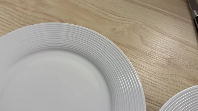 white ceramic plates with ribbed edges sit on a light brown wooden table, a metal knife and fork lie beside the tableware