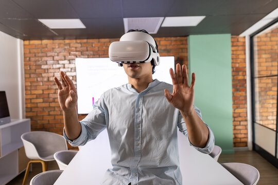 Asian man wearing white VR headset and light blue shirt, raising hands at conference table