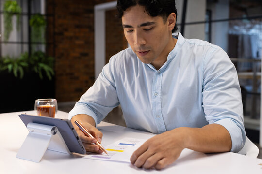 Asian man wearing light-blue shirt sleeves rolled sitting at white desk reviewing charts on tablet