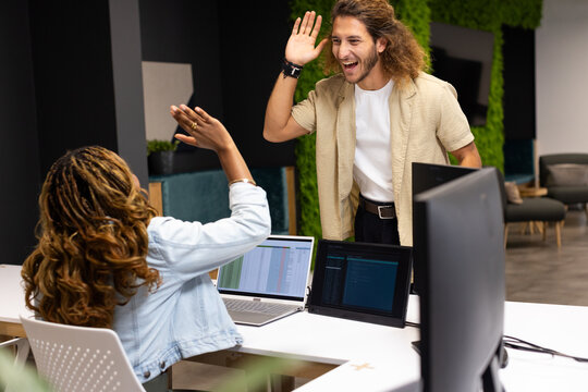 Diverse coworkers high-fiving while working at open-plan office with laptop, denim jacket
