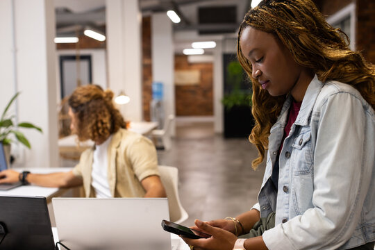 Diverse coworkers in open office, woman in denim jacket using phone, man using laptop, copy space