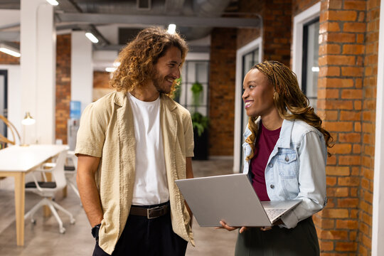Diverse coworkers chatting in open office while woman holding silver laptop, wearing denim jacket